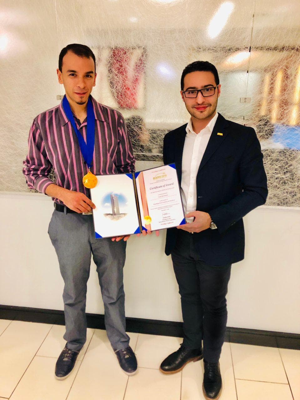 Un médaillé d’or tenant un certificat de prix BIXPO 2018, debout à côté d’un autre homme.