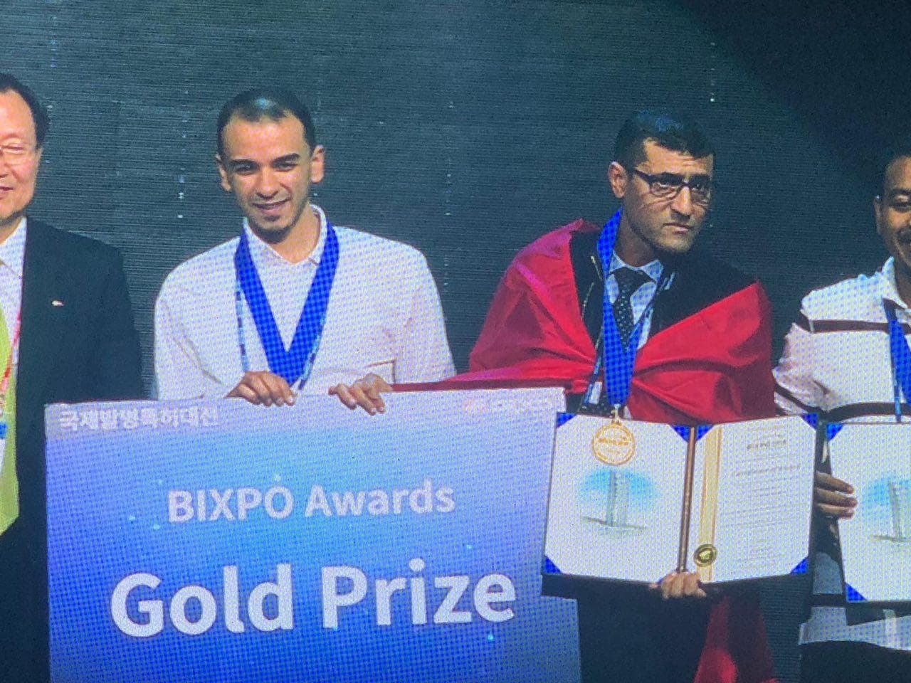 A presenter and medallists on stage at the BIXPO 2018 awards, behind a “BIXPO Awards · Gold Prize” board, one medallist holding an open award certificate.